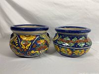 Two colorful Mexican ceramic pots side by side on white background, showing vibrant floral and leaf painting over blue glaze.