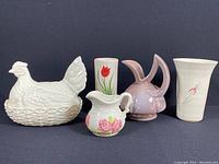 Five ceramic items arranged in a row: white chicken lidded dish, small floral pitcher, white vase with red tulip, Gonder pottery pitcher with dual spouts and handles, and white vase with small pink flower.