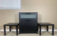 Front view of the black dresser with frosted glass drawer and two black side tables on each side against a wall with window blinds.