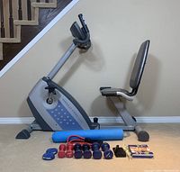 Photo showing Weslo Pro exercise bike with padded adjustable seat, blue exercise mat, various weights, resistance band, ankle weights, wrist weights, and fitness guide booklet arranged in front.