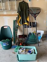 Full view of garden equipment lot including yard cart, rakes, shovels, waterproof poncho, broom, green hose, and container of gardening parts.