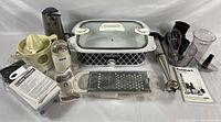 Full view of the lot including CrockPot, juicer attachment, laundry coffee grinder, electric can opener, Cuisinart hand blender and accessories laid out on a white surface