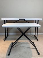 Side view showing both folding tables, larger table behind smaller one, both with white tops and black legs.