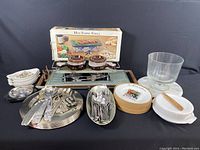 Wide shot showing the complete Hot Stone Grill with box, French onion bowls, metal charger plates stacked, utensils in trays, cutlery, and Brie maker with dessert glass dish beside.