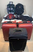 Full lot overview showing both red Heys suitcases front and center, with other soft bags and backpacks behind on table.