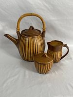 Photo shows the full tea set with teapot, creamer, and sugar holder, all ceramic with brown and yellowish vertical stripes, bamboo handle on teapot.