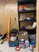 View of black plastic shelving unit with assorted tools and hardware in boxes and buckets alongside manual scraper tool.