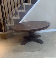 Round wooden pedestal table with clear plexiglass top placed in front of staircase.