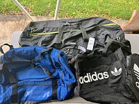 Photo of 3 duffle bags on a table outdoors: black Adidas bag, blue Hurley bag, and large black bag with yellow zipper pulls.