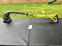 Full view of the Ryobi battery powered weedeater on a black table outdoors with grass background.