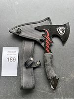 Camping tactical axe with black nylon sheath placed on dark surface with lot number tag