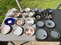 Overview of an outdoor table displaying an assortment of 19 vintage china pieces, including teacups and saucers with various floral and decorative patterns.