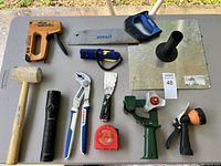Full overview of lot shows the staple gun, saw, mallet, flashlight, pliers, putty knife, tape measure, garden spray nozzle, plunger, and hose nozzle arranged on a table.
