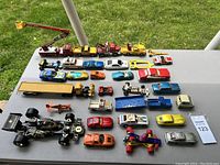 Photo showing the full lot of 31 toy cars and trucks arranged on a grey table outdoors with grass visible in the background. It highlights a diverse assortment of vehicles including race cars, construction trucks, and vintage cars.