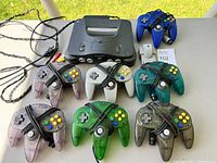 Wide shot of Nintendo 64 console and 7 controllers arranged on a table showing colors and condition.