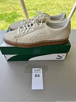 Side and profile view of off-white Puma sneakers on green and white Puma shoe box on outdoor table, showing stitching details on gum sole.