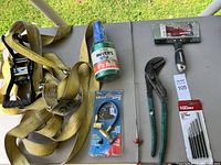 Top-down photo showing yellow ratchet straps, green wire spool, black-handled putty knife, blue-handled pliers, precision pick tool, packaged drill bit set, and propane torch adapter kit on a table