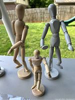 Four wooden artist mannequin models in natural and painted finishes arranged on a table outdoors with grass in the background, showing front and side views.