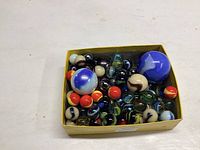 Top-down view of the box filled with assorted vintage glass marbles, mostly black, blue, red and white, with swirled and solid colors, in various sizes.