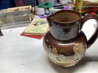 Front right-side view of brown glazed ceramic pitcher with raised white relief scenes of trees and figures.
