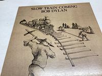 Front cover of the vinyl album 'Slow Train Coming' showing illustrated artwork of a man holding a cross with train tracks and a train in background.
