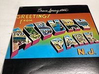 Front cover of Bruce Springsteen vinyl record 'Greetings From Asbury Park N.J.' showing colorful stylized lettering and images.