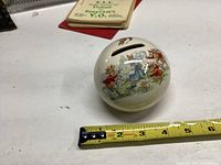 Round cream ceramic Bunnykins bank with painted rabbits celebrating, shown alongside measuring tape indicating about 4 inches diameter