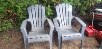 Two gray plastic lawn chairs placed outdoors on dirt ground next to bushes and garden tools.