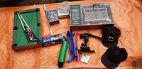 Photo showing a variety of items including mini pool table with balls and cues, foosball table set, speed stacking cups, skate guards, and two Batman hats arranged on orange fabric.