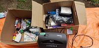 Two boxes of mixed office supplies and a black Fellowes Powershred paper shredder on an orange surface outdoors.
