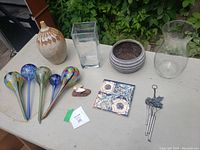 Various ceramic, glass vases, planter, decorative tile, four colorful glass water globes, and a metal wind chime on a table outdoors.