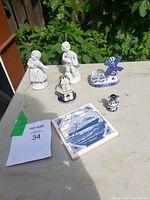 Overview of all items including boy and girl statues, two windmills, salt shaker, and coaster on a beige surface with green foliage behind.