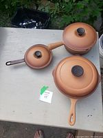 Three Le Creuset cookware items on a table outdoors: two pots with lids and one frying pan with lid, all in gradient orange to brown color, showing usage marks.