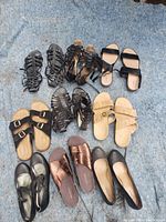 Top-down photo showing 11 pairs of assorted sandals and flats arranged on a blue fabric surface, including black strappy sandals, beige slide sandals, black flats, and metallic bronze slip-ons.