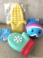 Three new dog toys displayed together on a couch: yellow corn-on-the-cob chew toy, blue-purple ball toy, teal mitten toy with red cuff and snowflake design.