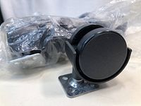 Side angle showing the black hard plastic double wheel caster fixed to a metal mounting plate, with the caster and a plastic bag behind it.