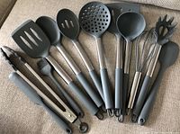 Set of 12 new silicone kitchen utensils laid out on a surface, showing black silicone heads and stainless steel handles with hanging loops.