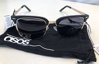 Front view of Asos sunglasses on a branded pouch showing black and gold frame detail with dark lenses.