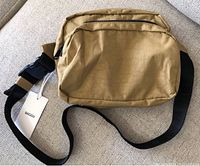 Front view of beige Baggu fanny pack showing main zippered compartment, smaller zippered pocket in front, attached black adjustable strap with plastic buckle, tag attached showing new condition.