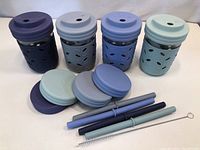 Set of four glass drinking cups each with a different blue silicone sleeve, lids, and straws with a cleaning brush