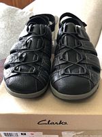 Front view of new black leather Clarks men's sandals placed on Clarks box, showing stitching and toe design.