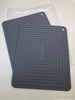 Two grey silicone trivets stacked, showing front and back surfaces with ridges and hanging holes, placed on white surface with packaging underneath.