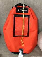 Front view of bright orange Sterling rope bag with black trim and clear front pocket and yellow drawstring cord tied at front.
