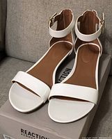 Front view of new white women's sandals on box showing cushioned tan footbed, ankle strap with gold buckle, and wide front strap.