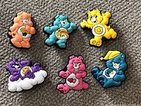 Photo showing all 6 Care Bear shoe charms laid out on a carpet background. Each charm features a different bear character with distinct colors and belly badges.