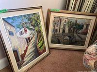 Two framed unsigned oil paintings leaning on carpeted floor showing architectural and natural scenes.