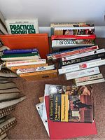 Photo showing a collection of hard and softcover cookbooks arranged on a carpeted floor, visible titles include Practical Problem Solving, various cookbook titles, and a boating book.
