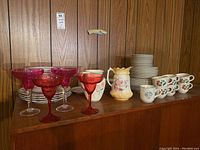 Wide shot showing entire assortment of dishes, pitchers and cocktail glasses on a wood shelf against wood paneling.