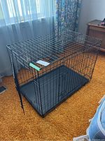 Front and side view of the collapsible metal dog crate with door open showing wire mesh and plastic tray bottom on carpet.