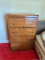 Front view of the wooden dresser showing four drawers with metal handles and knobs, placed beside a bed with red carpet flooring.